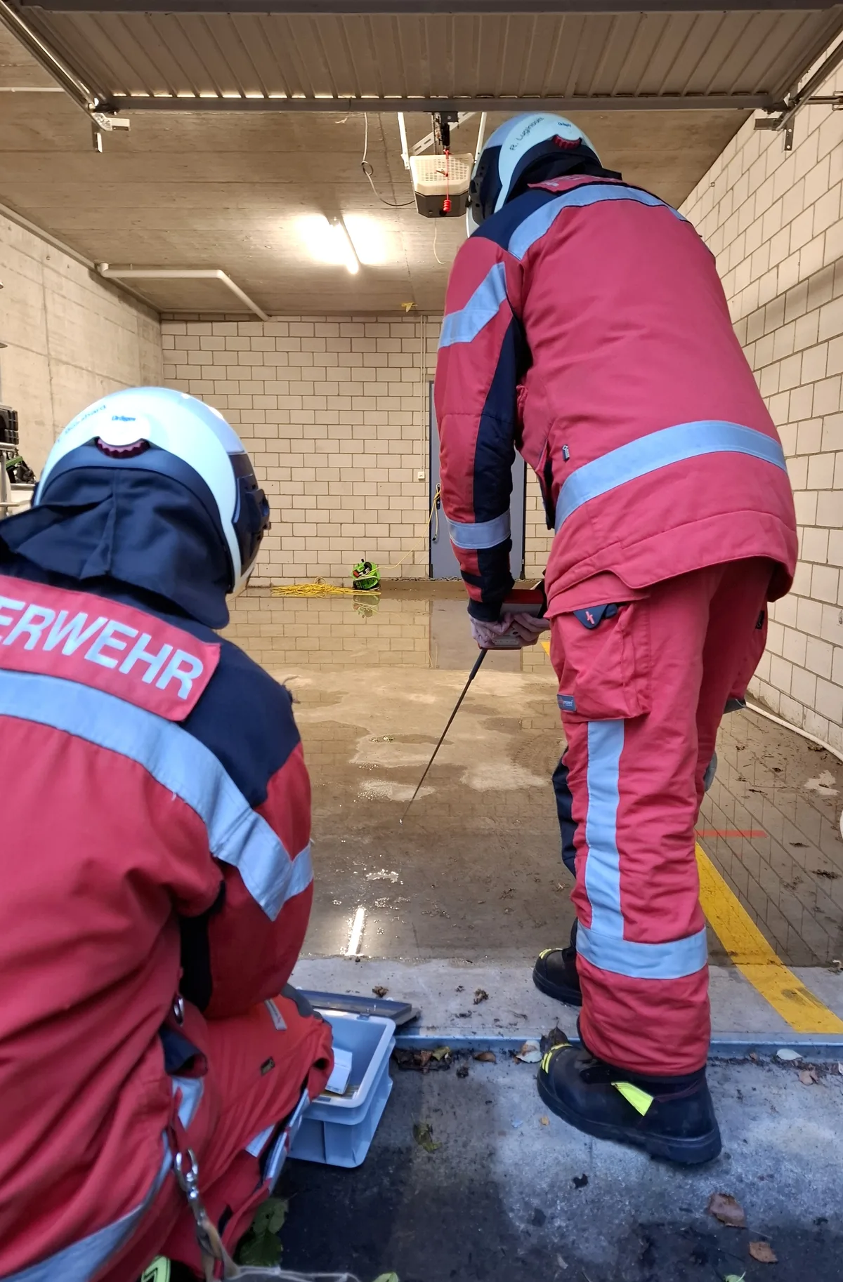 Die Feuerwehr überprüft, ob durch die Flüssigkeit auf dem Boden der Garage Strom fliesst. Zwei Feuerwehrleute stehen vor einer Garage, in der Wasser auf dem Boden zu sehen ist.
